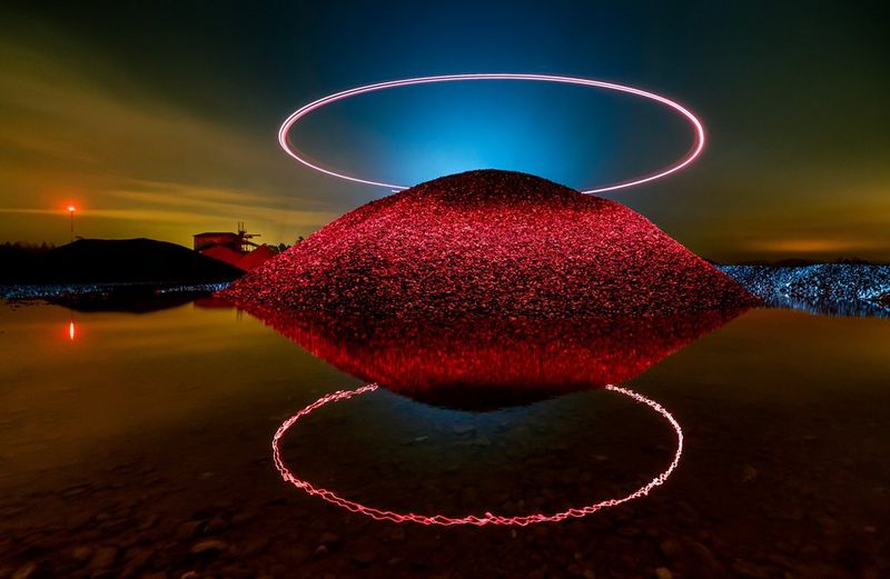 A mound of stone is illuminated by a halo of red light above it, which is also reflected in the water at its base.