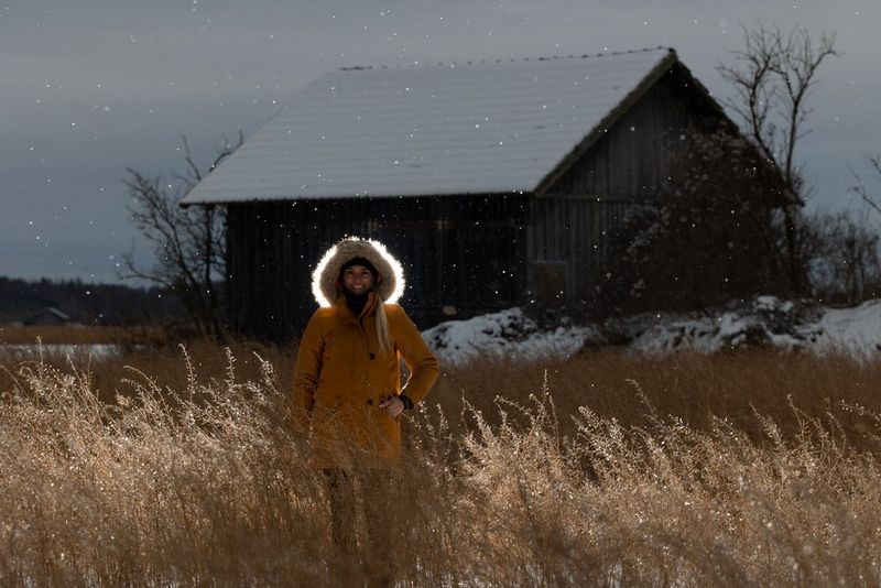 A backlit image of a woman standing in a field with long grass in the snow in front of an old wooden building.