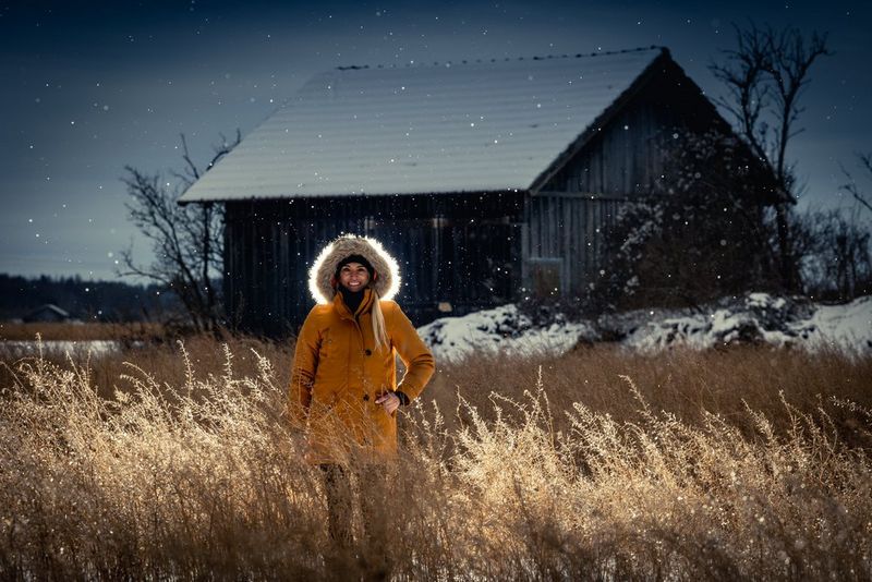 The same backlit image of a woman standing  in a field in front of a wooden building, edited to reveal more detail in the shadows. 
