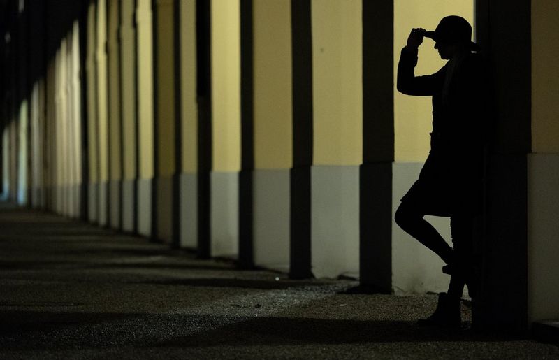 A silhouetted figure in a hat leaning against a wall beside a row of columns with light shining through them.