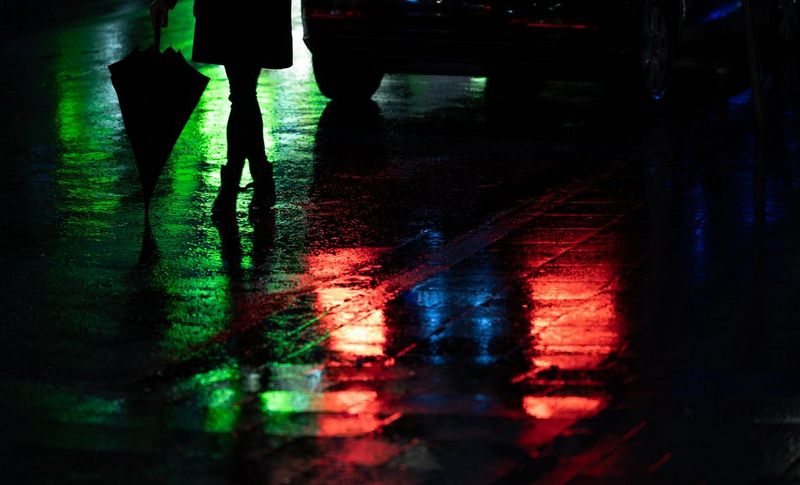 A figure visible waist-down holding an umbrella, standing with legs crossed on a rain-slick road beside a car, at night. Red light from the car's rear lights and green light from out of frame is reflected in the water.