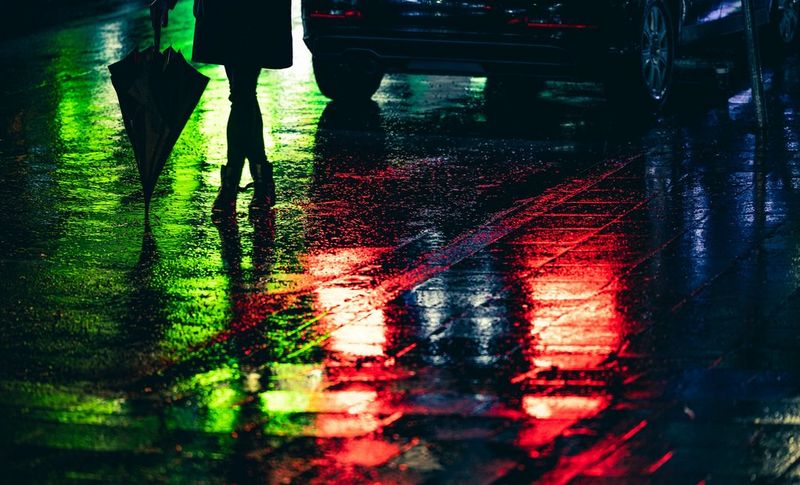 The same image of a figure holding an umbrella at night, standing with legs crossed on a rain-slick road beside a car, edited to brighten the colours and reveal detail in the shadows.