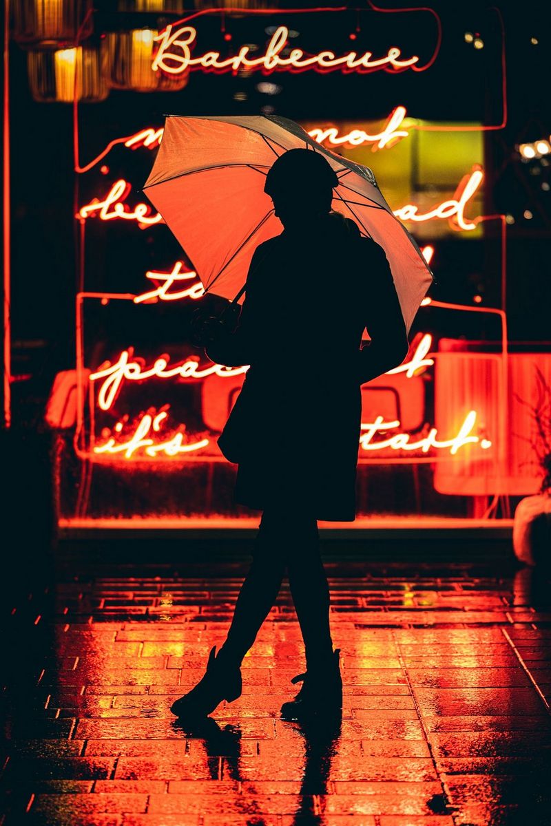 A silhouette of a woman with an umbrella standing outside a restaurant with an orange neon sign, the glow reflected in the wet pavement.