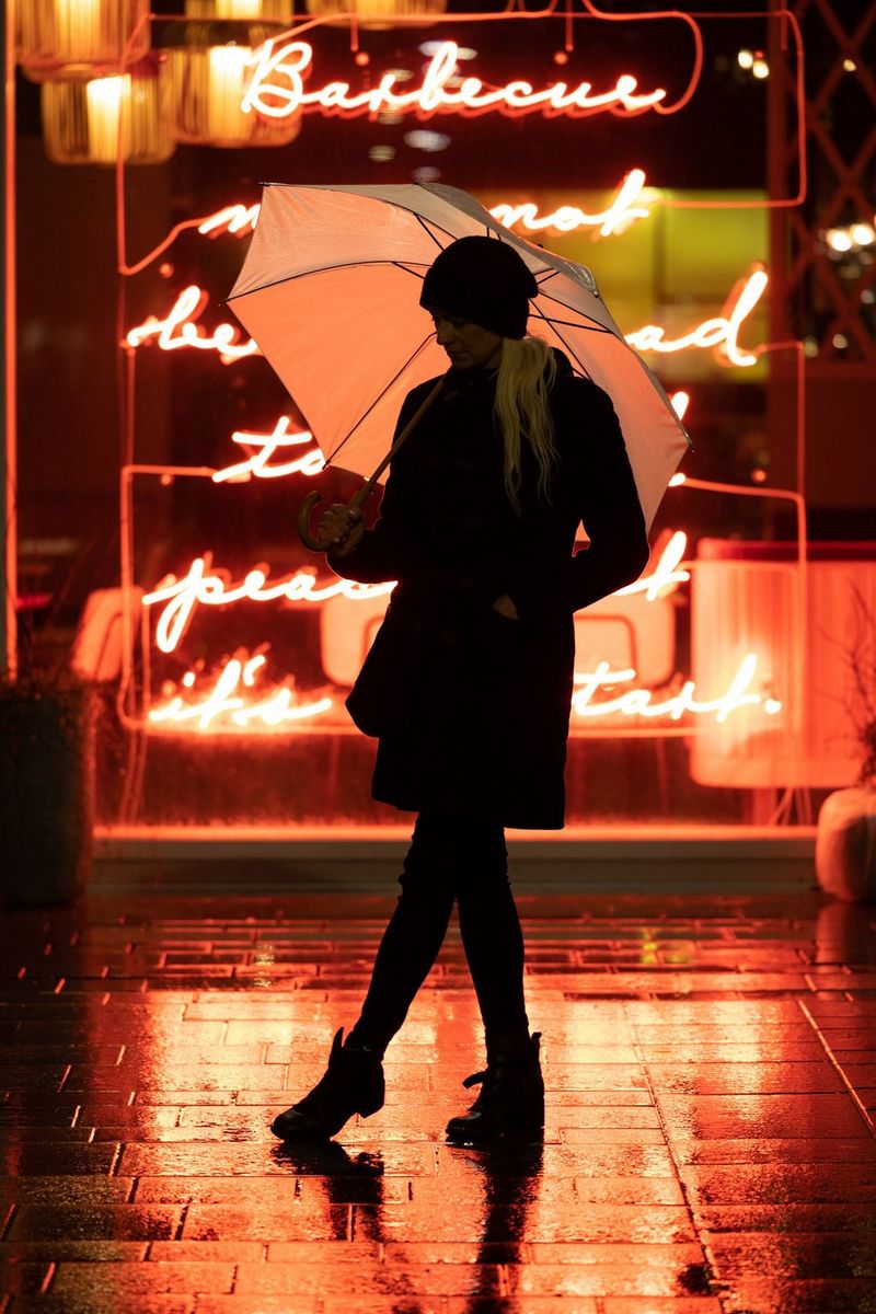 The same image of a woman with an umbrella standing outside a restaurant with an orange neon sign, edited so that more detail of her clothing and features is visible. 
