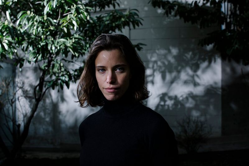 Een vrouw in een zwarte coltrui kijkt naar de camera, van opzij belicht en staande voor bomen vol bladeren die schaduwen werpen op de muur achter haar.