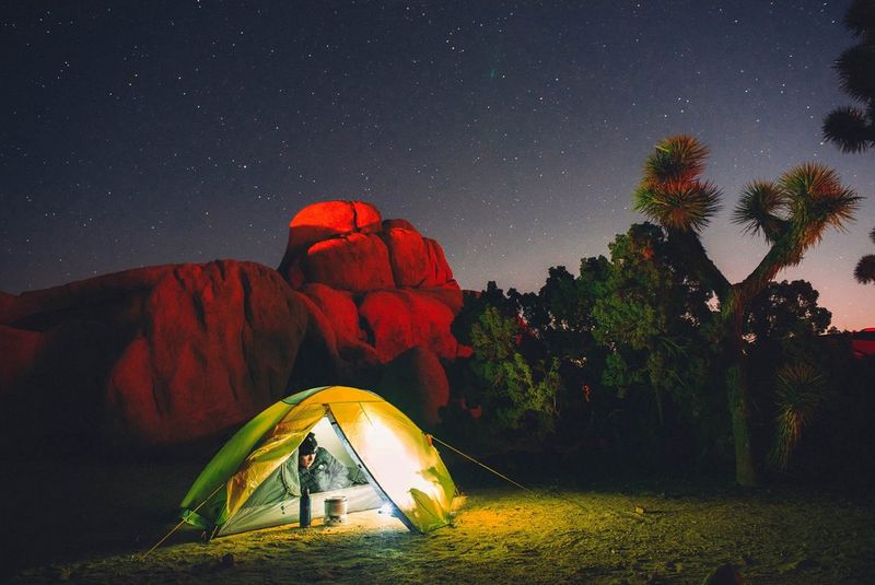 Een gele tent, verlicht door wit licht, staat op de voorgrond van rood verlichte rotsen, met een man in warme kleding die in de tent ligt en naar buiten kijkt.