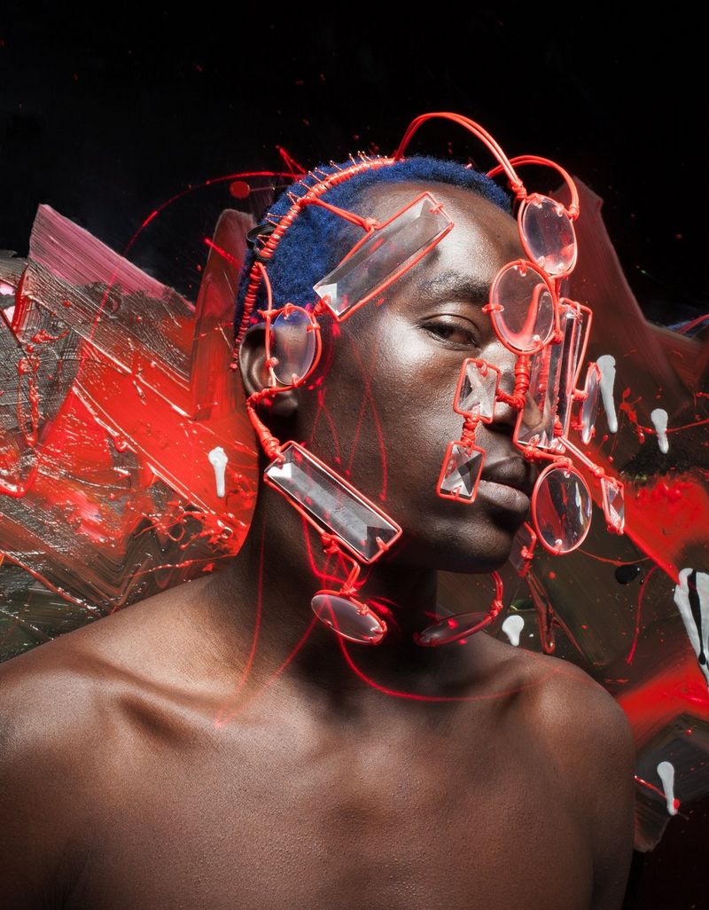Een man met ontbloot bovenlijf en blauw geverfd haar draagt een hoofddeksel van rode draad en doorzichtige lenzen, met lijnen van rood licht getekend rond zijn gezicht.