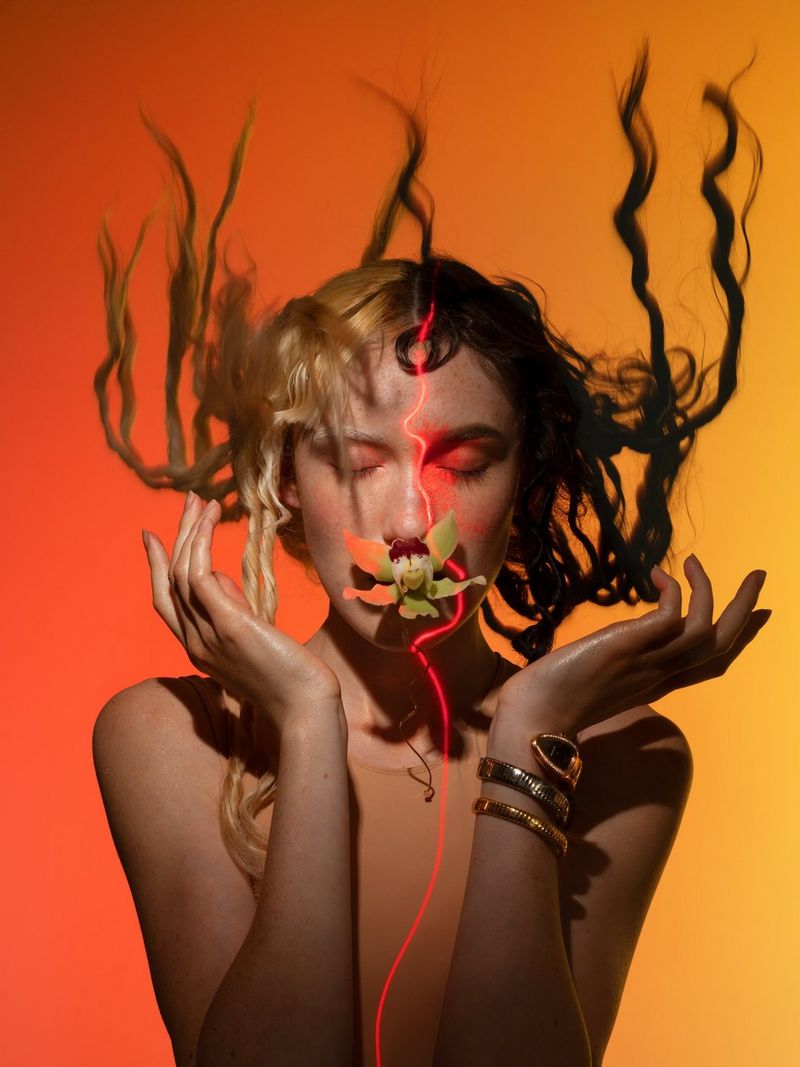 Een vrouw met aan de ene kant donker haar en aan de andere kant geblondeerd, houdt haar handen onder haar gezicht. Haar haren, in krullen gedraaid, steken boven haar hoofd uit, een open bloem bedekt haar mond en een lijn van rood licht loopt door het midden van haar haar en haar lichaam.