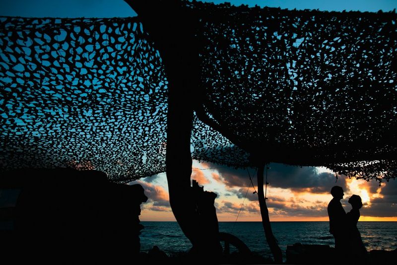 Een bruid en bruidegom staan afgetekend tegen een zonsondergang onder een donkere gaasluifel.