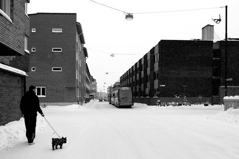 Een zwart-witfoto van een man die een hond uitlaat in een besneeuwde straat, terwijl in de verte een snowboarder een stunt uitvoert op het dak van een gebouw.