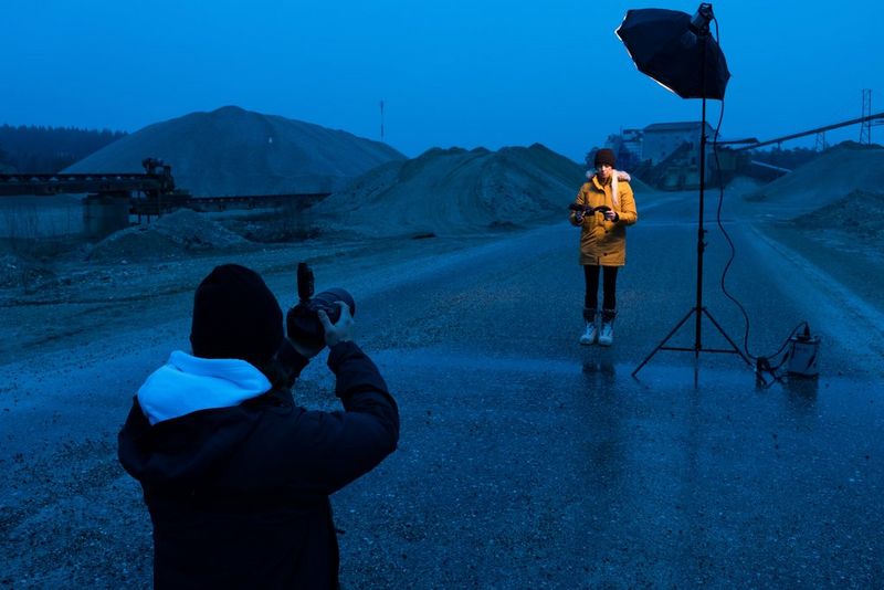 Un uomo fotografa una donna in piedi che indossa un giaccone giallo. È illuminata da una luce esterna; sullo sfondo un paesaggio industriale dai toni scuri.