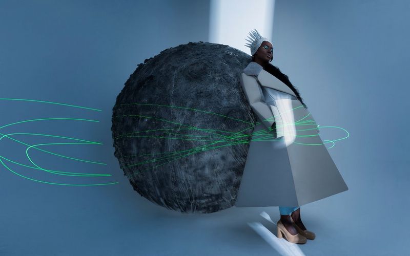 Immagine con esposizione lunga di una donna che indossa una corona e un lungo cappotto grigio, appoggiata a una grande palla grigia. In primo piano, delle scie di luce verde.