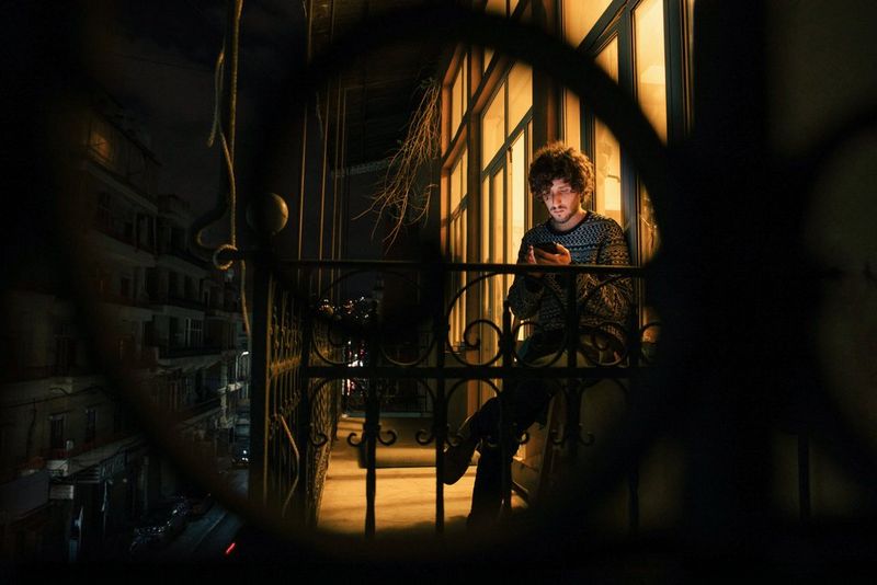 Un uomo seduto in un balcone in ferro battuto con lo sguardo rivolto al proprio telefono. È illuminato da una calda luce arancione proveniente dall'ambiente alle sue spalle.