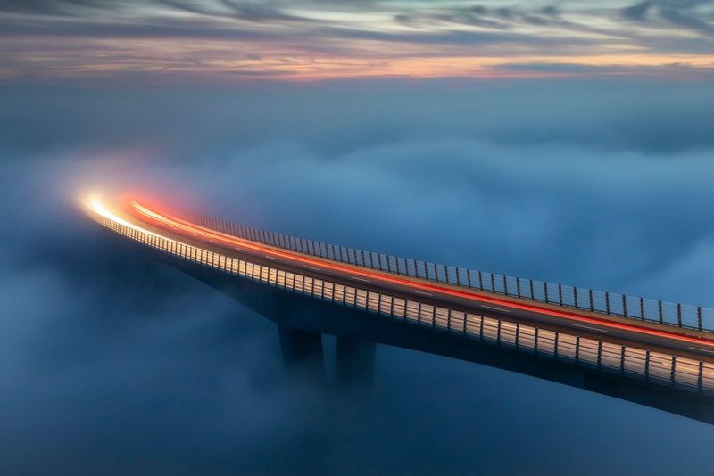 Traînées de lumière de voitures dans la lueur du soir à travers un pont incurvé, ses supports et le sol en dessous complètement obscurcis par un épais brouillard semblable à un nuage.