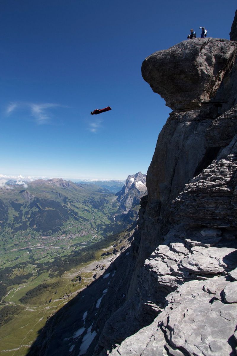 A figure in a wingsuit leaps into the abyss in the Swiss Alps as two crew members film from the clifftop.  