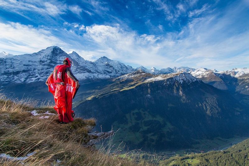 A man standing in a wingsuit on a mountain outcrop in the Swiss Alps. Several snow-capped peaks can be seen in the distance behind him. 