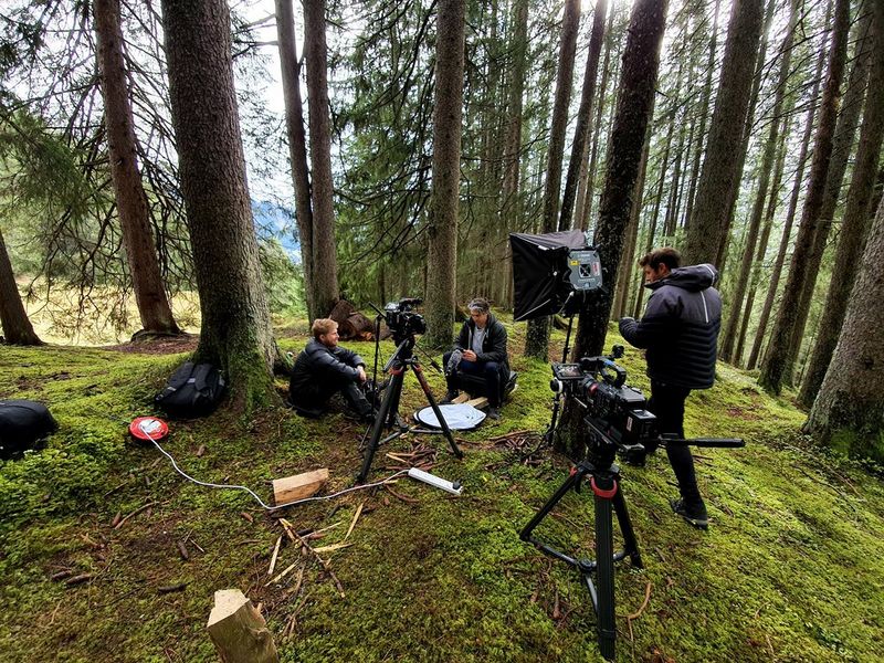 Three men gathered among the trees filming an interview, with a lightbox and Canon camera set up on a tripod. 