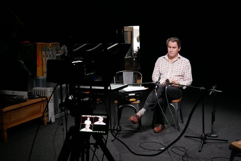 A man sits alone in a darkened studio with multiple views of him visible on the monitor attached to the camera in front of him.
