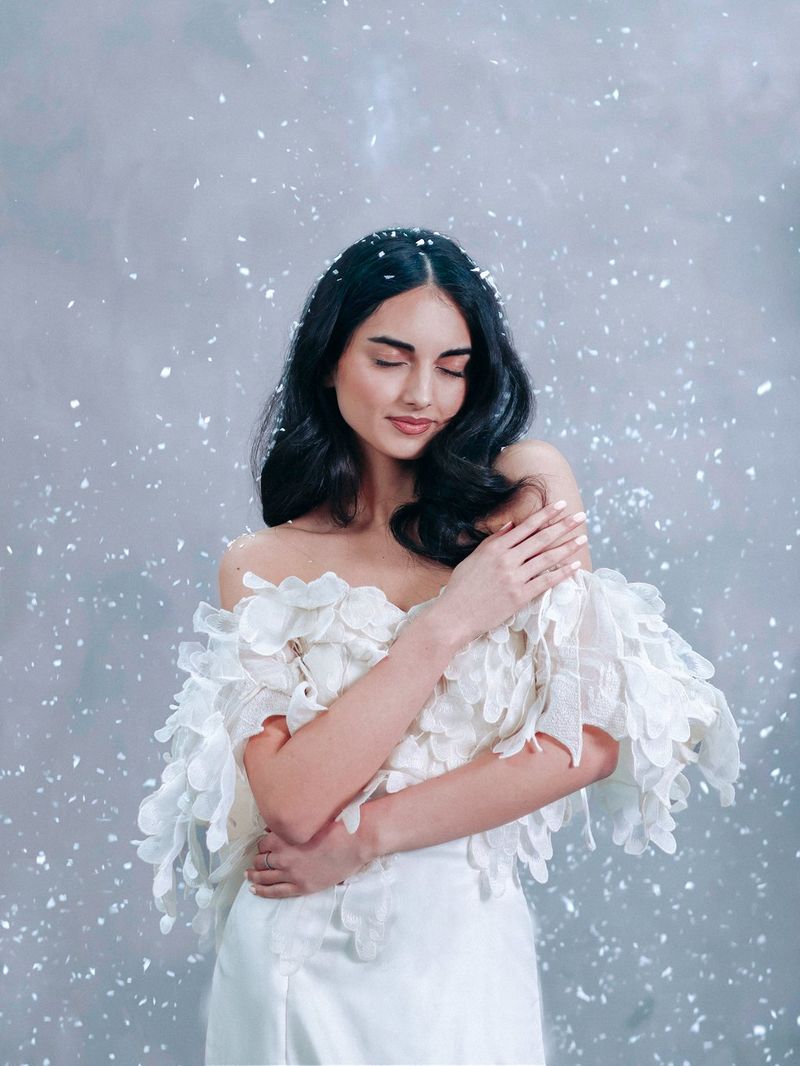 A woman in a white, off-the-shoulder dress with a feathered top holds her arms around her body, looking down to her left, as snowflakes appear to fall around her. Photograph by Rosie Hardy.