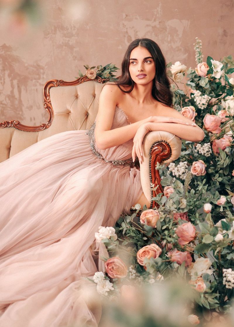 A woman in a light pink gown lies on a chaise longue, leaning against the arm, with white and pink flowers cascading next to her. Photograph by Rosie Hardy.