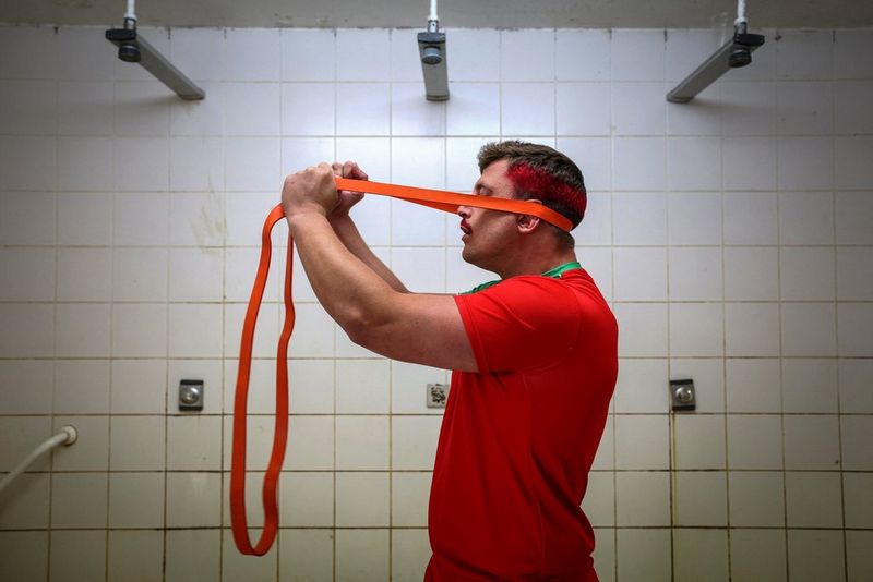 Giocatore di rugby nello spogliatoio, vicino alle docce, con indosso una maglia rossa e con i capelli neri con una striscia rossa, che scalda il collo con un elastico arancione poggiato dietro alla testa. Scatto realizzato con Canon EOS R3 e obiettivo Canon RF 24-70mm F2.8 L IS USM da Valentine Chapuis.