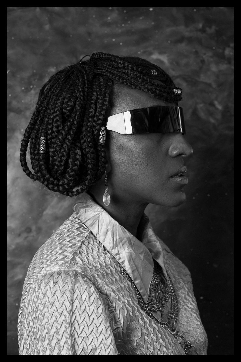 A black and white portrait of Sarah Waiswa, with wrap-around sunglasses and braided hair.