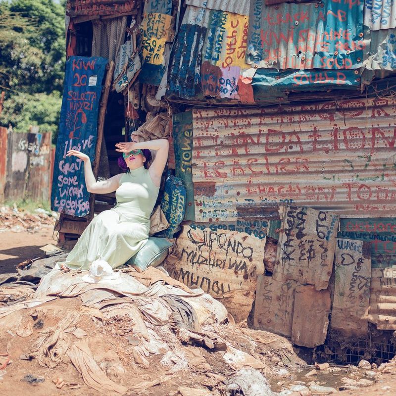 A pale-skinned woman in a long dress, sitting on a cushion at the door of a shanty with colourfully painted corrugated iron panels, raises her arms as if against the sun.