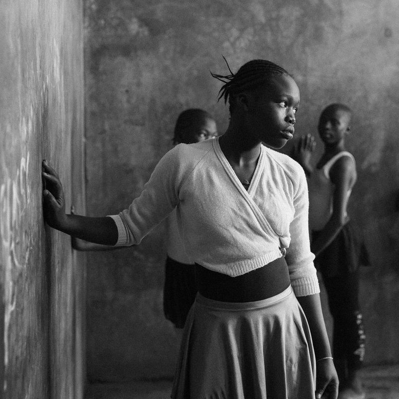 A young woman looks wistfully to the side, one hand on a wall. Two other people stand against another wall in the background.