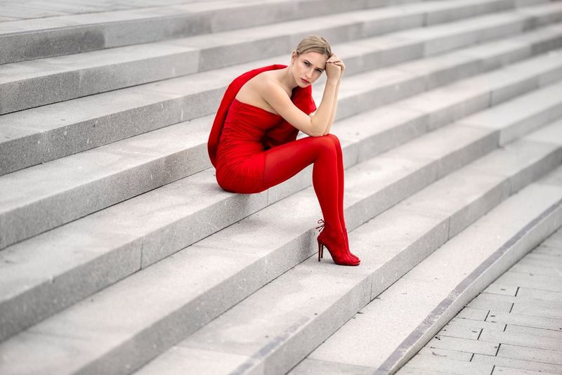 A woman wearing a red top, red bottoms and red heels sits on some steps and looks directly at the camera. Taken by Sascha Hüttenhain with a Canon RF 135mm F1.8L IS USM lens.
