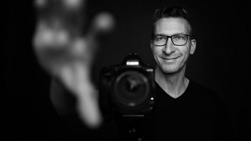 A smiling Sascha Hüttenhain extends his hand towards the camera used to photograph him in this black and white image. 