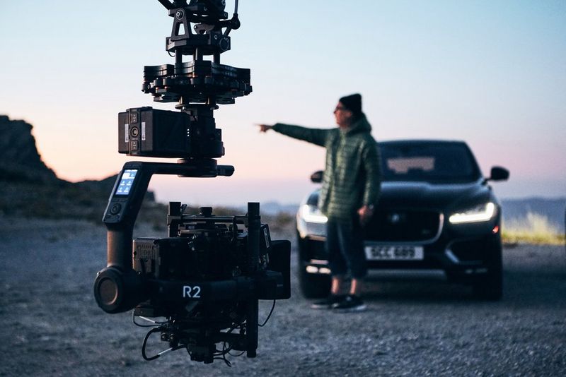 A person in a coat and hat stands in front of a car and points with their arm up, a Canon camera setup in the foreground.