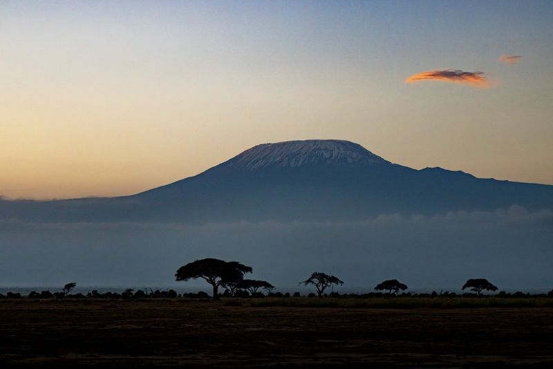 A flat-topped mountain is seen in the background, with a few trees silhouetted in the foreground and two small clouds in the top-right sky.