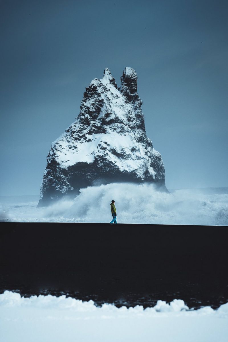 A figure walks along a black sand beach, in front of a large, snow-covered rock.