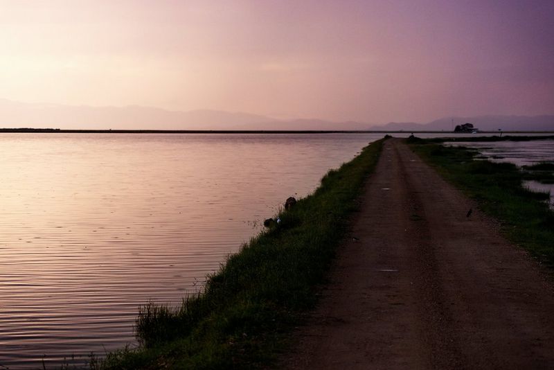 A dirt road with grass verges stretches out into the distance leading away from the camera, next to an expanse of rippling water.