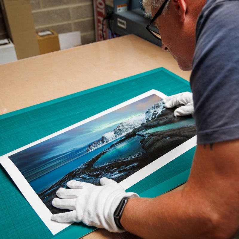 A person wearing white gloves and glasses examines a print of a wintry landscape with the Northern Lights in the sky above.