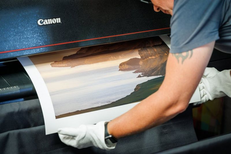 A man wearing white gloves leans forward to take a large landscape print from a Canon printer. 