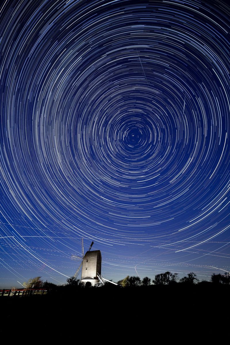 A night sky filled with circular star trails above an old-fashioned windmill. Several meteors cross the star trails in the top right of the picture.
