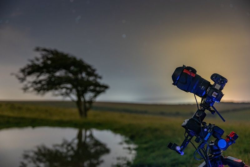 A Canon EOS R5 camera and lens are positioned on a tripod, tilted to face up at the sky, in front of a field, pond and windswept tree.