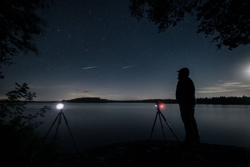 A man wearing a hat and warm clothes is silhouetted against a lake and night sky, next to two cameras on tripods pointing upwards. Two meteors cross the sky above him.