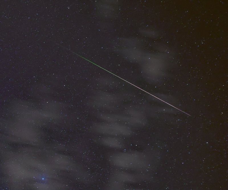 A single meteor crosses the sky, in front of several clouds. The line of the meteor is purple at the front, changing to green at the back.