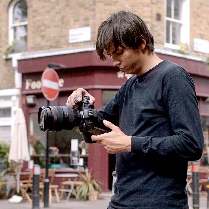 A young man dressed in black stands on a street corner, holding a Canon EOS R5 camera at chest height and looking down into its LCD screen.