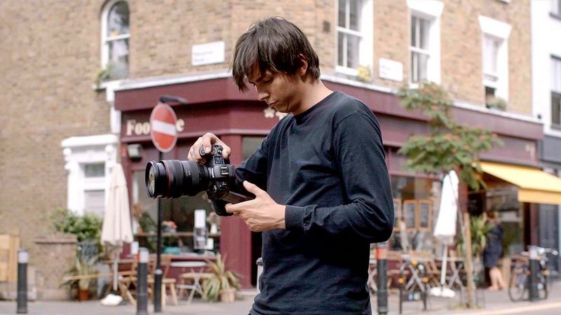 Een man in het zwart staat op de hoek van een straat. Hij heeft een Canon EOS R5-camera in zijn hand en houdt deze op borsthoogte. Hij kijkt omlaag naar het LCD-scherm van de camera.