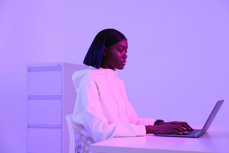 An image shot on a Canon EOS R5 by Jaroslav Monchak showing a model wearing a large white robe, sitting at a desk with a filing cabinet behind her and working on a laptop. The whole image is bathed in violet light. 