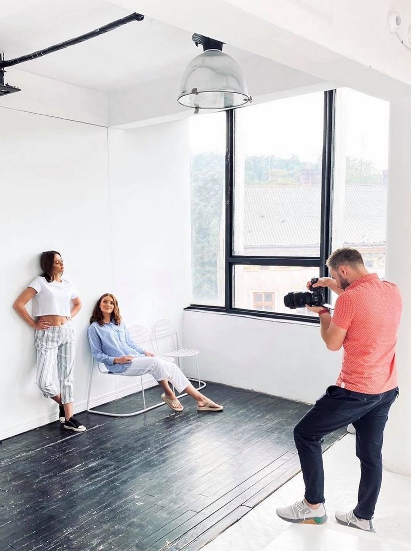 Fashion photographer Jaroslav Monchak shoots two women standing and sitting against a white wall, the whole room lit by the large window at the side.