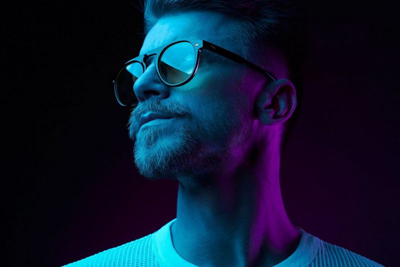 A close-up of a model's face, turned to the side. He has a neat moustache and beard and is wearing dark glasses. The background is dark, and the model is lit with blue and purple light.