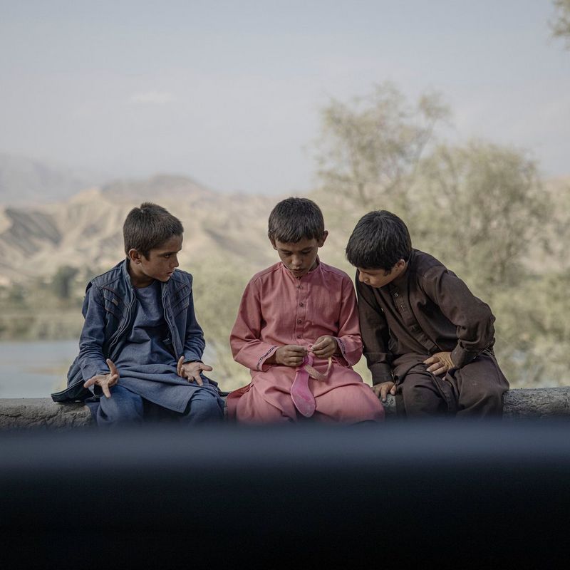 Three young Afghan boys deep in discussion sit on a wall in front of a large body of water. One of the boys is holding a bright pink sandal.