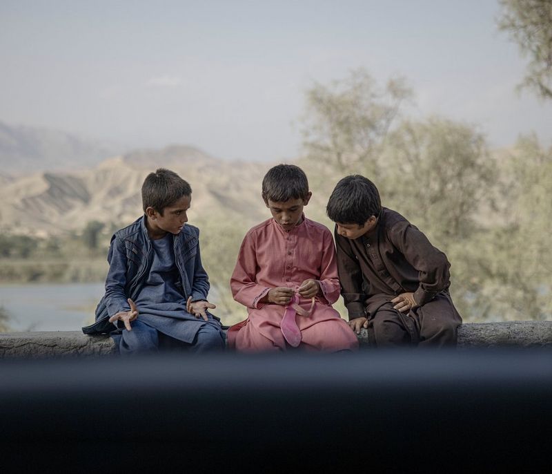 Три афганских мальчика разговаривают, сидя на стене перед большим водоемом. Один из мальчиков держит ярко-розовую сандалию.