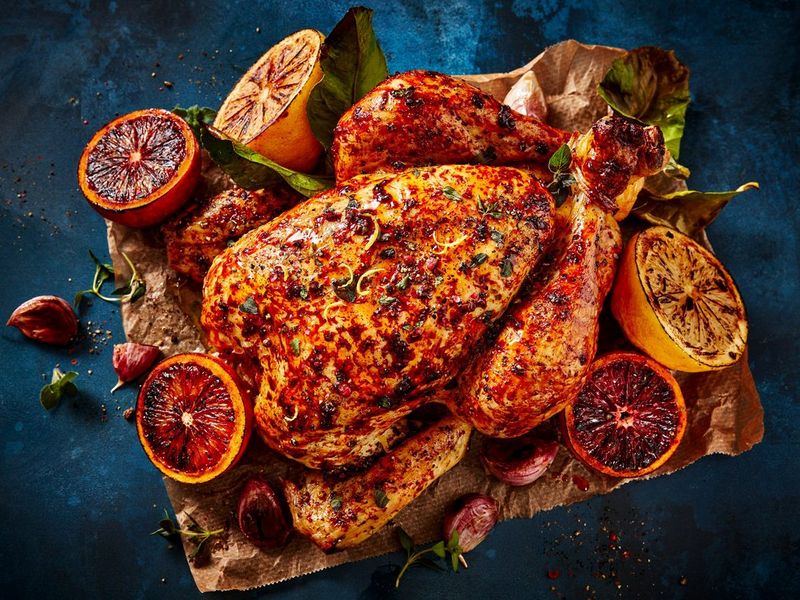  A roast chicken sits on some brown paper on a dark blue table. It's covered in herbs and spices, and surrounded by halves of oranges and cooked garlic cloves.