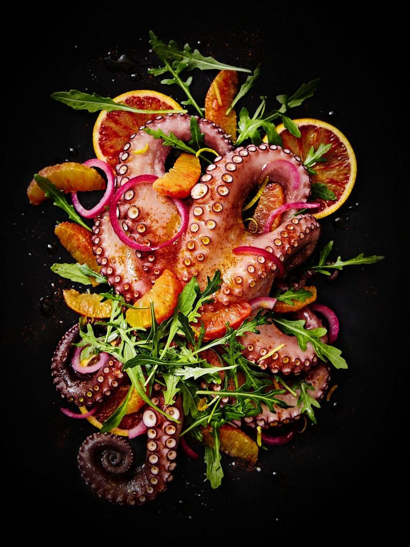 A cooked octopus in shades of pink, purple and orange sits on a black background, covered with segments of blood orange and bright green leaves.