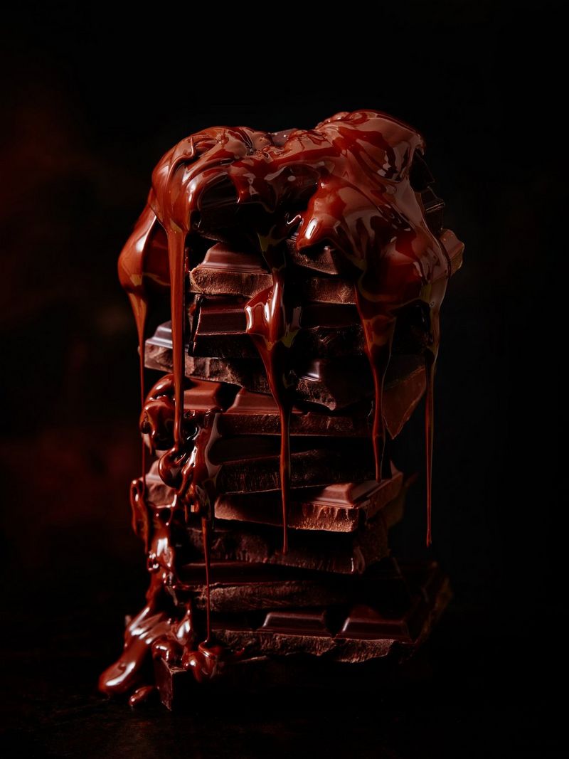A stack of broken up bars of chocolate dripping with melted ganache set against a black background.